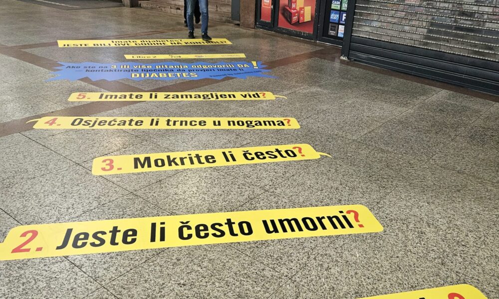 Fotografija prikazuje unutrašnjost javnog prostora, vjerojatno željezničke ili autobusne stanice. Na podu su postavljeni veliki žuti natpisi s pitanjima na hrvatskom jeziku, koji se odnose na simptome dijabetesa. Pitanja su numerirana od 1 do 5 i glase: Pijete li puno vode? Jeste li često umorni? Mokrite li često? Osjećate li trnce u nogama? Imate li zamagljen vid? Ovi natpisi vode prema plavom natpisu koji sugerira da bi odgovori na ova pitanja mogli ukazivati na dijabetes. U pozadini se vide stepenice i pokretne stepenice, kao i nekoliko plakata na zidovima. Desno se nalazi zatvoreni kiosk s natpisom "TISAK".