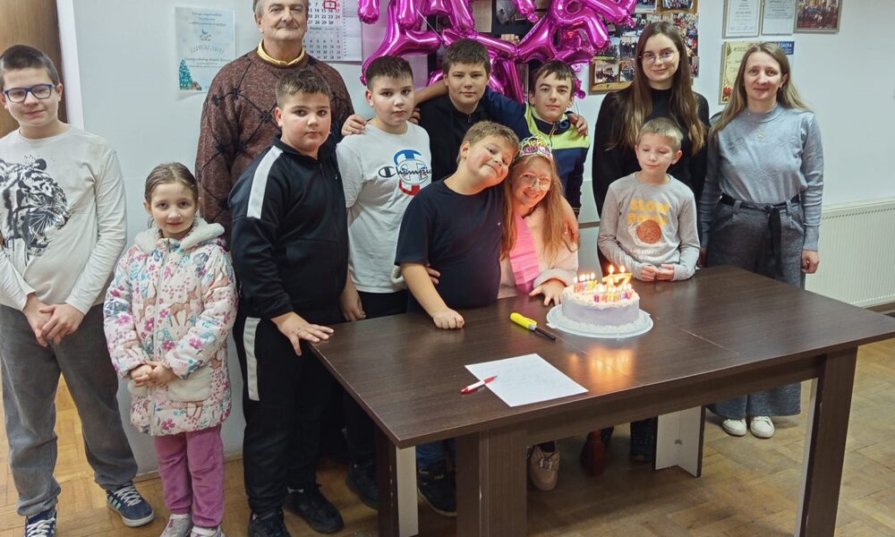 Na fotografiji je grupa ljudi koja stoji oko stola na kojem se nalazi torta s upaljenim svjećicama. Iza njih su baloni koji tvore natpis "HAPPY BIRTHDAY". Na zidu iza njih nalaze se razni plakati i kalendari. Na stolu se također nalazi papir i olovka.