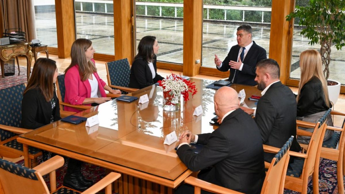 Na fotografiji je prikazan sastanak u uredu. Sedam osoba sjedi oko velikog drvenog stola s prozirnom staklenom površinom. Na stolu se nalazi cvjetni aranžman u sredini, a ispred svake osobe su papiri i olovke. U pozadini se vide veliki prozori kroz koje se pruža pogled na vanjski prostor s drvećem i stazama. Sastanak se odvija u formalnom okruženju, a sudionici su odjeveni u poslovnu odjeću. Na stolu su također postavljene pločice s imenima sudionika.