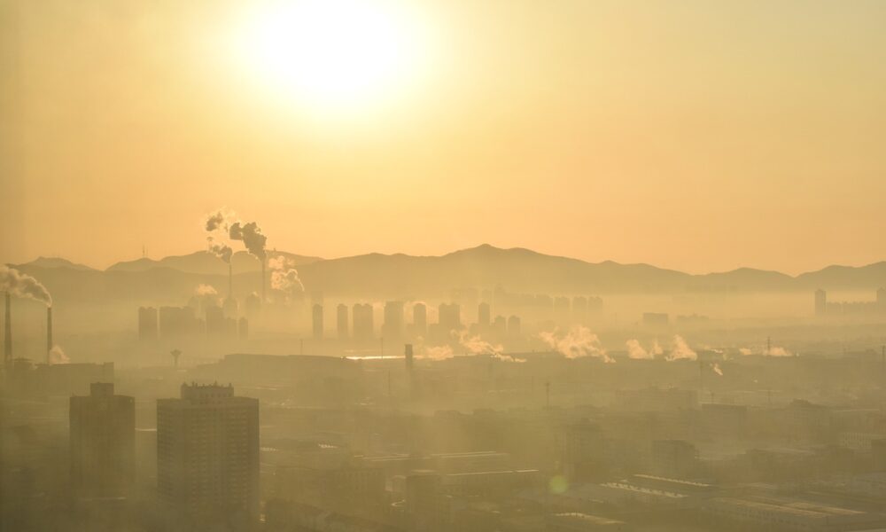 Fotografija prikazuje urbani krajolik pod zlatnim svjetlom izlazećeg ili zalazećeg sunca, obavijen gustim slojem smoga. U daljini se vide dimnjaci i industrijski kompleksi koji ispuštaju dim, dok se iza njih naziru obrisi visokih zgrada i planinskog lanca. Maglovita atmosfera stvara efekt zamagljenosti, naglašavajući problem zagađenja zraka u urbanim sredinama.