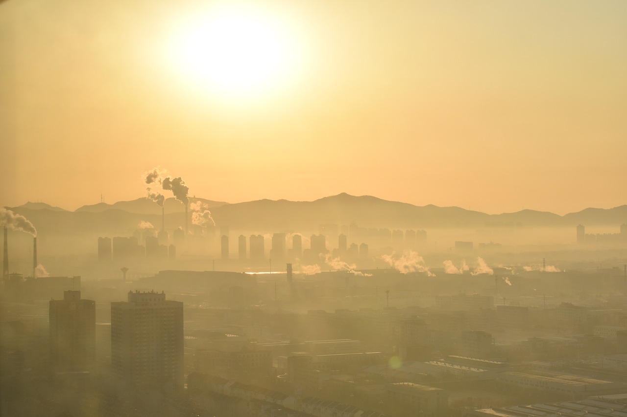 Fotografija prikazuje urbani krajolik pod zlatnim svjetlom izlazećeg ili zalazećeg sunca, obavijen gustim slojem smoga. U daljini se vide dimnjaci i industrijski kompleksi koji ispuštaju dim, dok se iza njih naziru obrisi visokih zgrada i planinskog lanca. Maglovita atmosfera stvara efekt zamagljenosti, naglašavajući problem zagađenja zraka u urbanim sredinama.