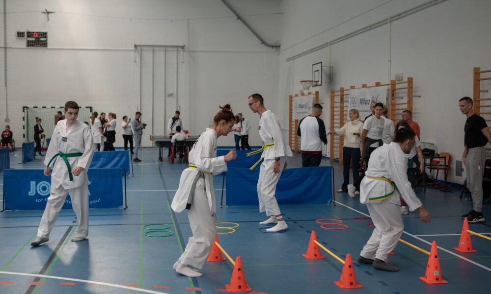 Fotografija prikazuje više djece kako treniraju u bijelim kimonima u sportskoj dvorani. Na fotografiji se vide plave strunjače i rekviziti za vježbanje koje osobe preskaču.