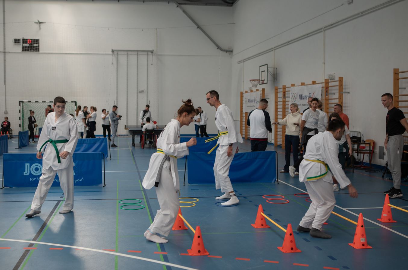 Fotografija prikazuje više djece kako treniraju u bijelim kimonima u sportskoj dvorani. Na fotografiji se vide plave strunjače i rekviziti za vježbanje koje osobe preskaču.