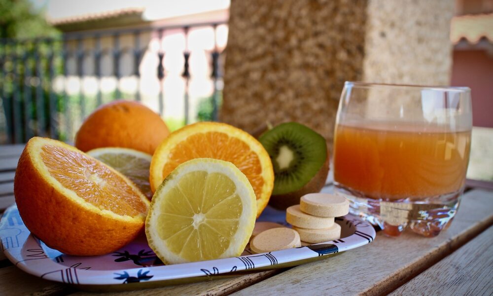 Fotografija prikazuje tanjur s narezanim voćem koje uključuje naranče, limun i kivi, te čašu soka od naranče. Pored toga, na stolu se nalaze i nekoliko tableta ili dodataka prehrani. Scena je postavljena vani, vjerojatno na terasi ili balkonu s pogledom na zid od opeke i drveće u pozadini. Ova fotografija može biti zanimljiva ili relevantna zbog fokusa na zdrav životni stil i važnost vitamina iz voća.