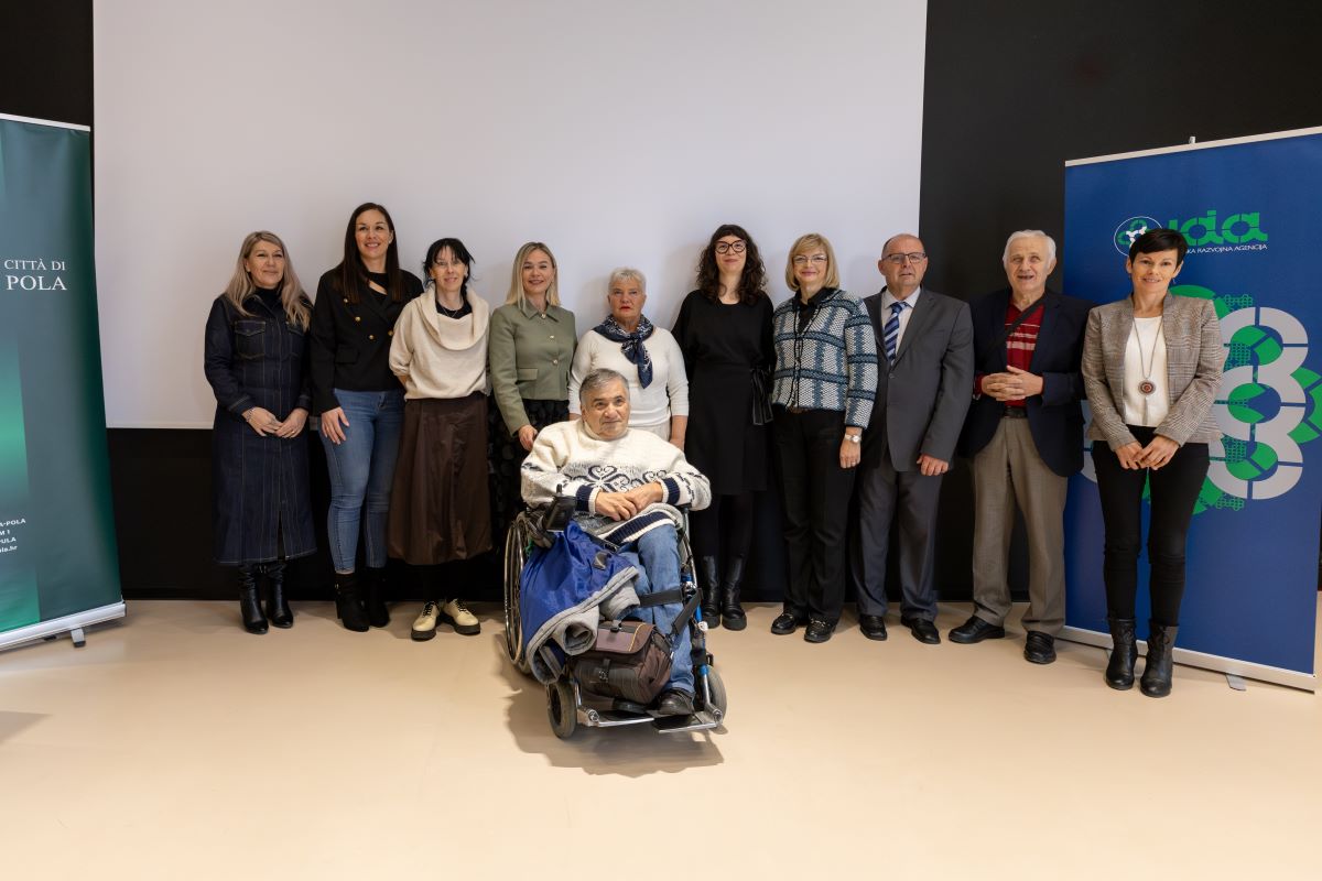 Na fotografiji je grupa od jedanaest osoba koje stoje ispred bijelog platna. Jedna osoba u prvom planu sjedi u invalidskim kolicima. Svi su obučeni u poslovnu ili ležernu odjeću. S lijeve strane fotografije nalazi se zeleni plakat s natpisom "Città di Pola", dok se s desne strane nalazi plavi plakat s logotipom i natpisom "Istarska razvojna agencija". Fotografija izgleda kao da je snimljena tijekom nekog službenog događaja ili sastanka.