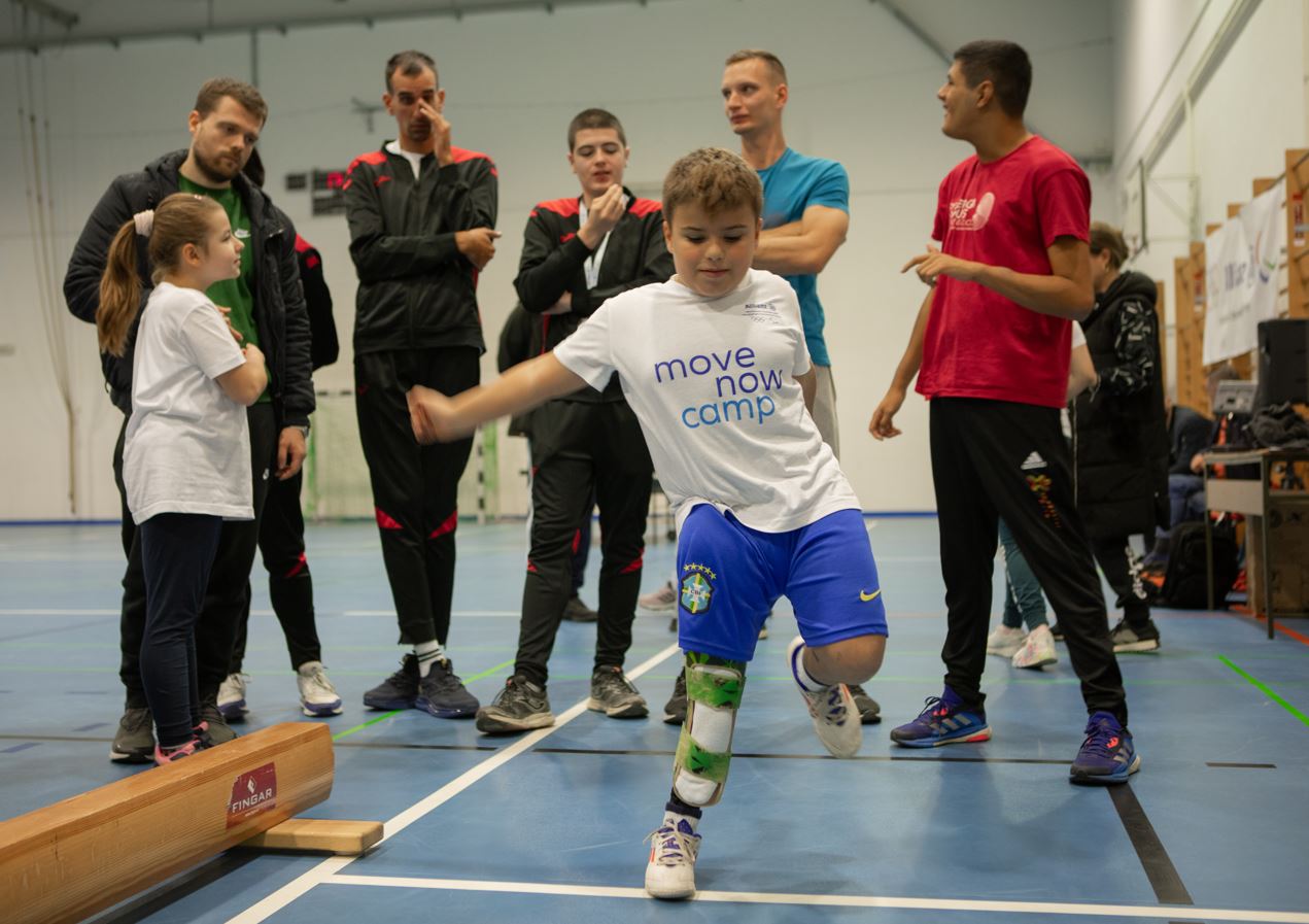 Fotografija prikazuje nekoliko ljudi, a između ostalog jednog dječaka sa protezom na jednoj nozi koji pokušava šutnuti nešto.