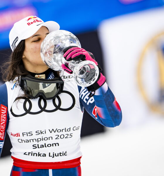 Skijašica Zrinka Ljutić stoji u skijaškom odijelu s hrvatskim obilježjima i bijelom kapom s logom Red Bull, dok ljubi veliki kristalni pehar. Na bijelom prsluku koji nosi piše: Audi FIS Ski World Cup Champion 2025 – Slalom – Zrinka Ljutić. U pozadini se vide zastave i sportski transparenti. Atmosfera je svečana i pobjednička.