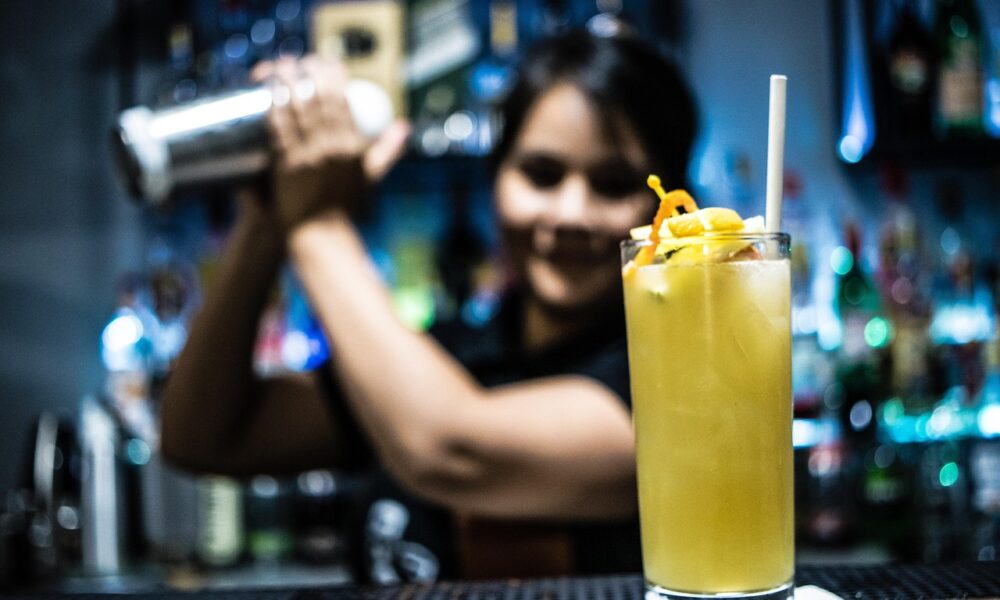 Fokusirana slika žutog koktela u visokoj čaši s ledom, slamkom i ukrasom od narančine kore i limuna, postavljenog na bijelu papirnatu salvetu na šanku. U pozadini, zamućena figura barmenice koja se smije i drži shaker, pripremajući piće. Ambijent je noćni bar s policama punim raznih boca, osvijetljenih plavičastim svjetlom.
