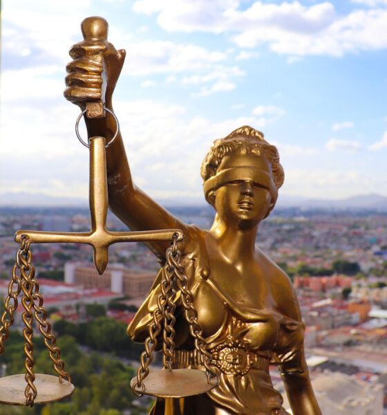 Zlatna statua božice pravde Justicije s povezom preko očiju, drži vagu u podignutoj ruci. Vaga ima dva tanjura obješena na lancima, simbolizirajući ravnotežu i pravičnost. U pozadini se kroz stakleni prozor pruža panoramski pogled na grad s mnoštvom zgrada, zelenilom i planinama u daljini pod vedrim nebom s oblacima.