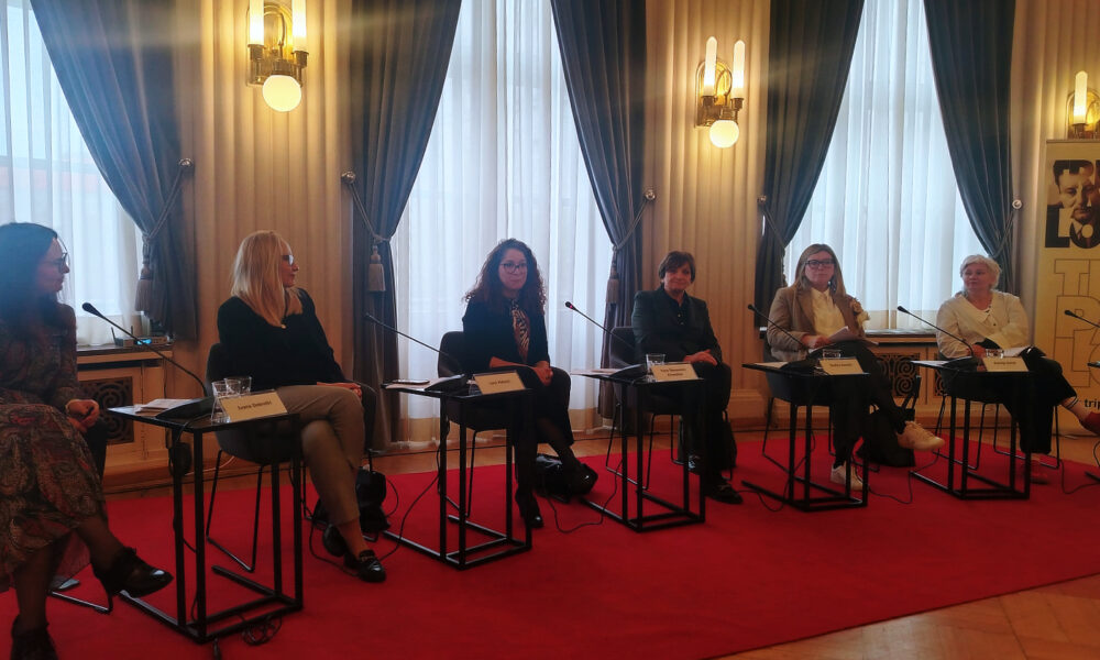 Šest žena sjedi u redu iza mikrofona tijekom panel-rasprave u elegantnoj prostoriji s crvenim tepihom i visokim prozorima prekrivenim tamnim zavjesama. Ispred svake govornice nalaze se čaše vode i pločice s imenima. U pozadini se vidi plakat Centra za demokraciju i pravo Miko Tripalo.