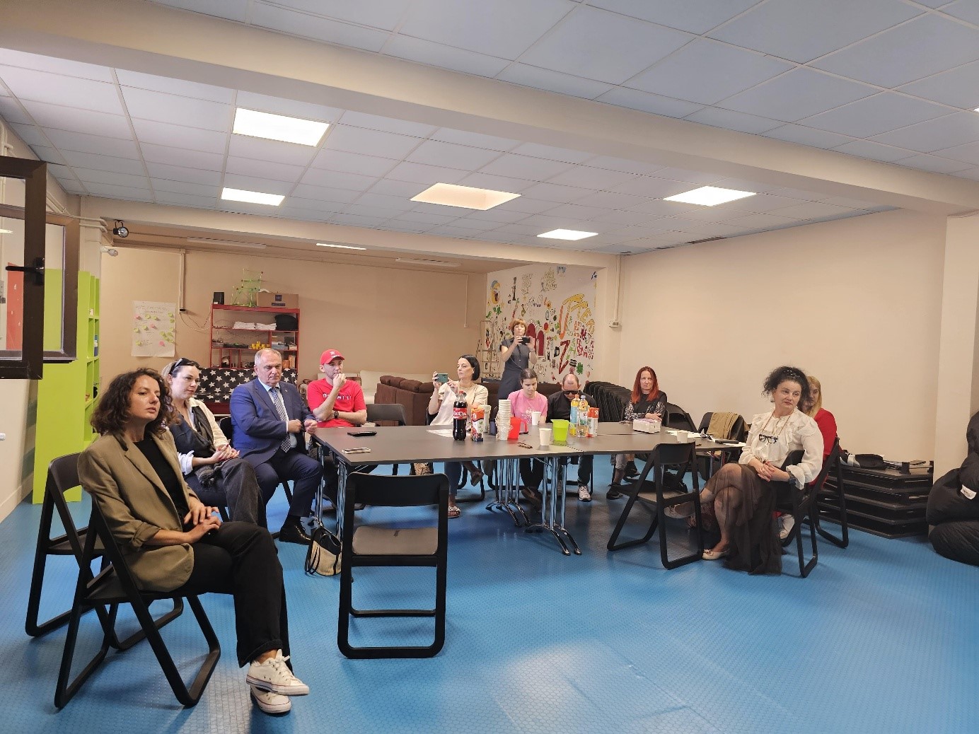 Na fotografiji je prikazana skupina ljudi okupljenih u zatvorenom prostoru, najvjerojatnije dvorani ili višenamjenskoj prostoriji. Osobe sjede oko pravokutnog stola i u njegovoj blizini, pažljivo prateći događaj, što sugerira da je riječ o sastanku, radionici ili okruglom stolu. Na stolu se nalaze pića i grickalice, što dodatno upućuje na neformalnu i ugodnu atmosferu. U prostoru se vidi plavi gumirani pod, što je karakteristično za prostore koji se koriste za različite aktivnosti. Zid u pozadini ukrašen je šarenim crtežima, što prostoru daje vedrinu. Na fotografiji su prisutne osobe različite dobi i stila odijevanja, a u prvom planu s lijeve strane sjedi žena u bež sako-u i bijelim tenisicama. Među okupljenima se nalazi i muškarac u odijelu, što upućuje na prisutnost predstavnika institucija ili organizatora događaja. Cjelokupan dojam fotografije je pozitivan i prikazuje okupljanje u inkluzivnom i poticajnom okruženju, vjerojatno u sklopu Programa DANAS.