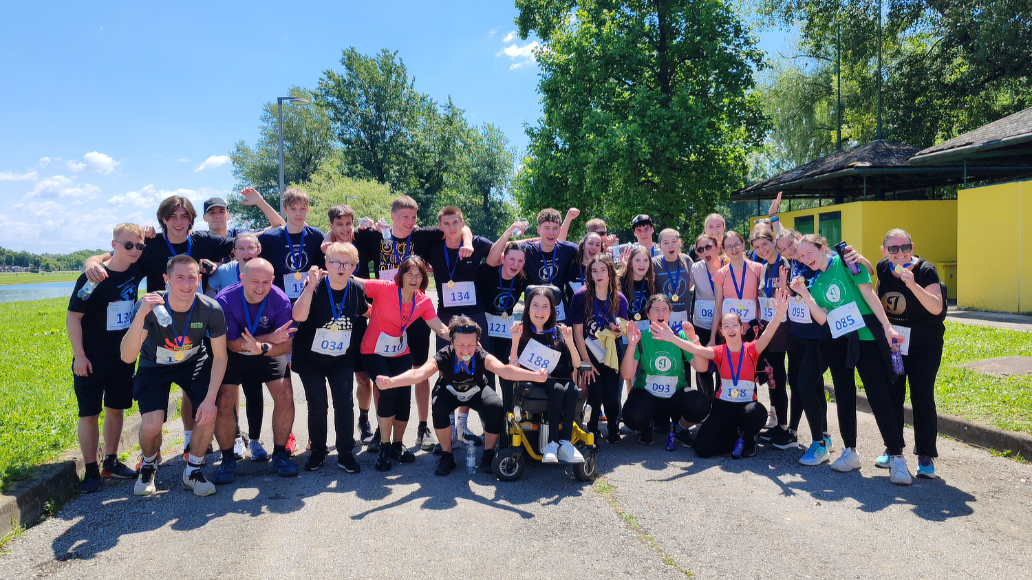Velika skupina mladih i odraslih osoba okupljena na stazi uz jezero, svi nasmijani i s medaljama oko vrata. Neki drže boce vode, a nekoliko osoba u prvom planu pozira s podignutim rukama. Među njima su i osobe u invalidskim kolicima. Svi imaju startne brojeve, što sugerira da je riječ o inkluzivnoj sportskoj manifestaciji ili humanitarnoj utrci. Sunčano vrijeme i zelenilo pojačavaju dojam pozitivne i svečane atmosfere.