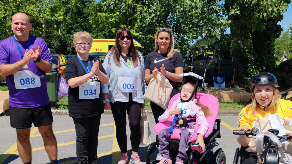 Grupa šestero ljudi stoji nasmijana na otvorenom, ispred drveća i ambulantnog vozila. Nose medalje i startne brojeve, sugerirajući sudjelovanje u sportskom događaju. Među njima je djevojčica u invalidskim kolicima, kao i žena na ručnom biciklu s kacigom na glavi. Svi poziraju i plješću, u atmosferi zajedništva i veselja.