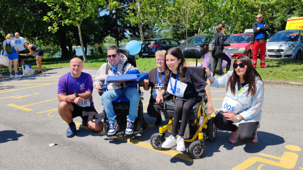 Pet osoba pozira nasmiješeno na parkiralištu u blizini livade i drveća. Dvije osobe koriste električna invalidska kolica i nose medalje oko vrata, kao i ostali sudionici. Svi nose startne brojeve. U pozadini se nalaze automobili, ljudi i vozilo hitne pomoći. Atmosfera je prijateljska i podržavajuća.