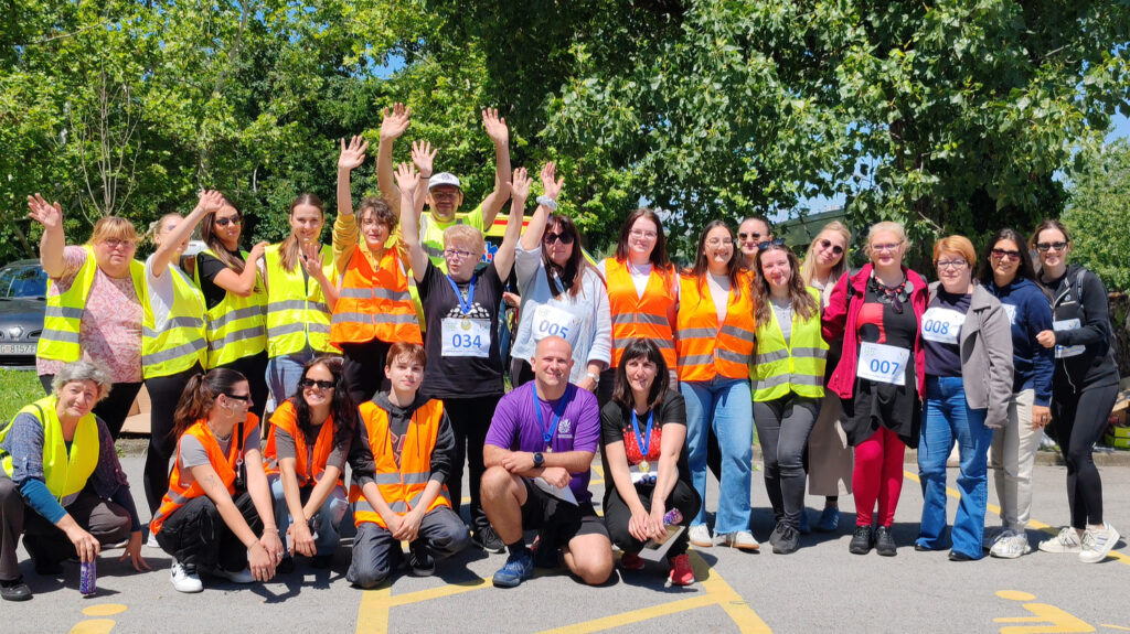 Grupna fotografija velikog broja volontera i sudionika sportskog događanja, od kojih mnogi nose reflektirajuće prsluke u narančastoj i žutoj boji. U prvom planu sjedi nekoliko mladih osoba, a iza njih stoje stariji i mlađi sudionici koji poziraju nasmiješeni, s podignutim rukama. Okruženi su drvećem i prirodom, a dan je sunčan i vedar.