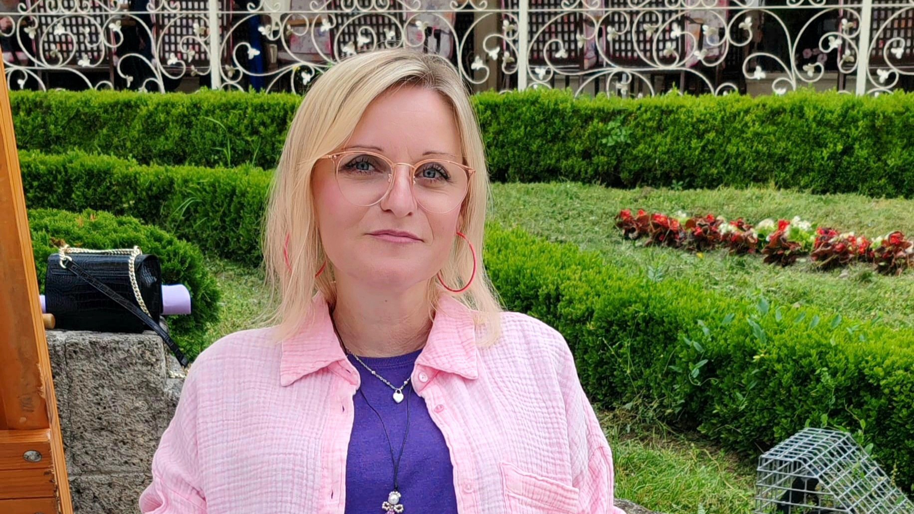 Na fotografiji se vidi osoba koja sjedi na stolici na otvorenom. Osoba nosi ružičastu košulju preko ljubičaste majice, uz plave traperice. Oko vrata ima ogrlicu s privjeskom, a na desnoj ruci tetovažu ruže. U pozadini je lijepo uređeni vrt s niskim zelenim grmljem, cvijećem i metalnom ogradom. S lijeve strane slike nalazi se drveni stalak, a iza osobe je crna torba.