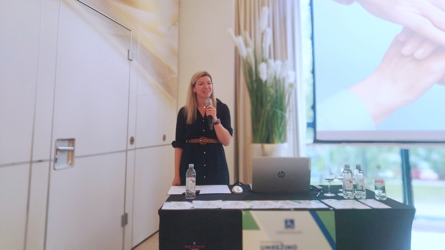 Žena stoji iza stola i drži mikrofon, obraćajući se publici na konferenciji. Na stolu su prijenosno računalo, boce vode i papiri. U pozadini se nalazi veliki ekran s prikazom ruku koje se dodiruju i visoka vaza s bijelim cvijećem. Atmosfera je profesionalna i svečana.
