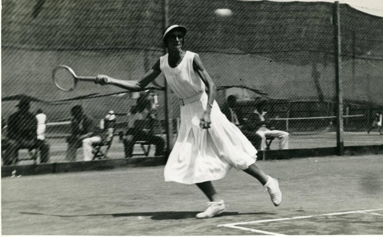 Crno bijela fotografija prikazuje tenisačicu obučenu u suknju kako igra tenis.