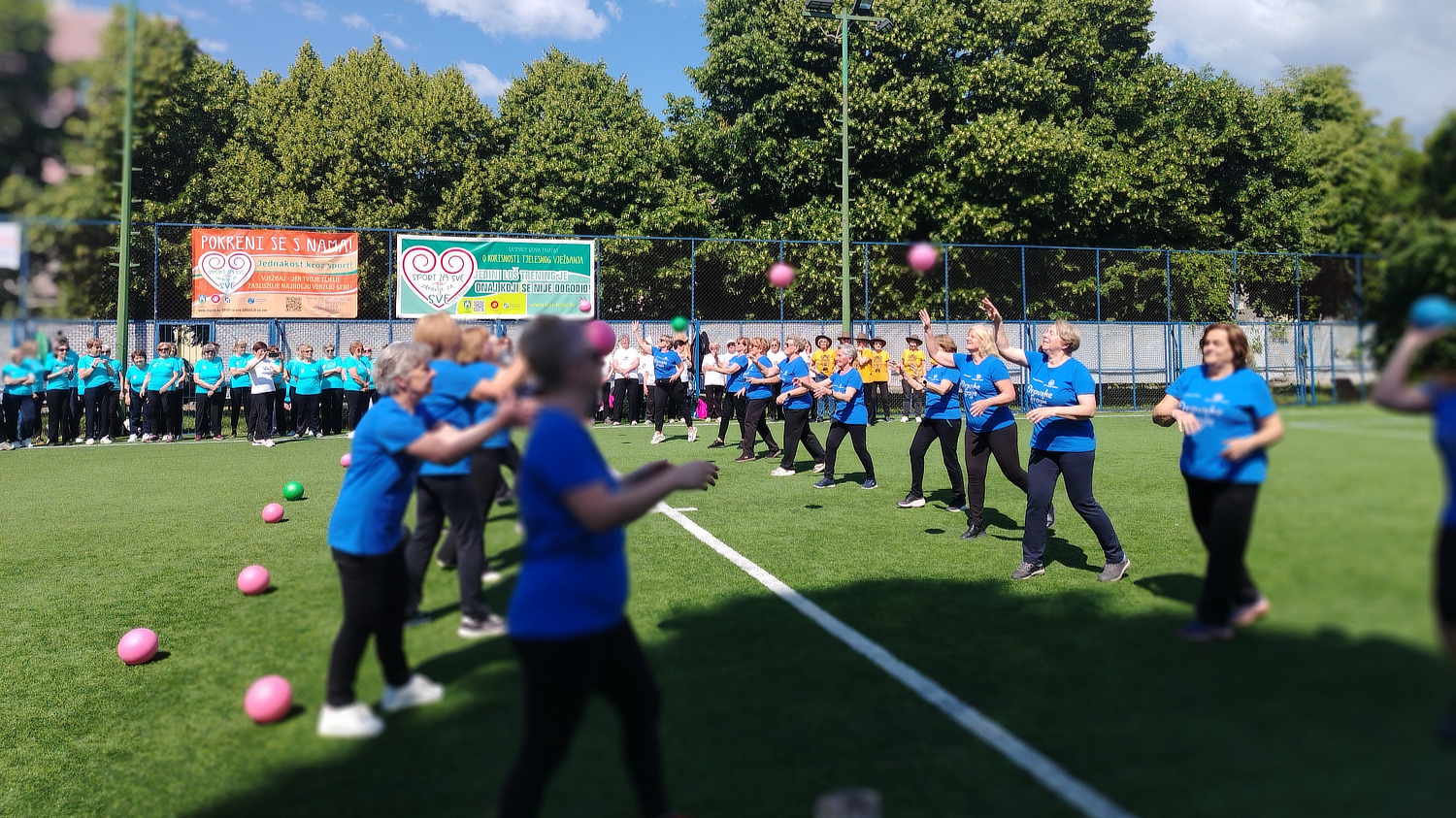 Grupa starijih žena u plavim majicama vježba na sportskom terenu na otvorenom, bacajući i hvatajući šarene lopte. U pozadini se vidi više grupa ljudi u različitim majicama, koji stoje uz ogradu, promatraju ili čekaju svoj red. Na ogradi su istaknuti transparenti s natpisima poput „Pokreni se s nama“ i „Sport za sve“. Okruženje je zeleno i sunčano, s drvećem u pozadini.