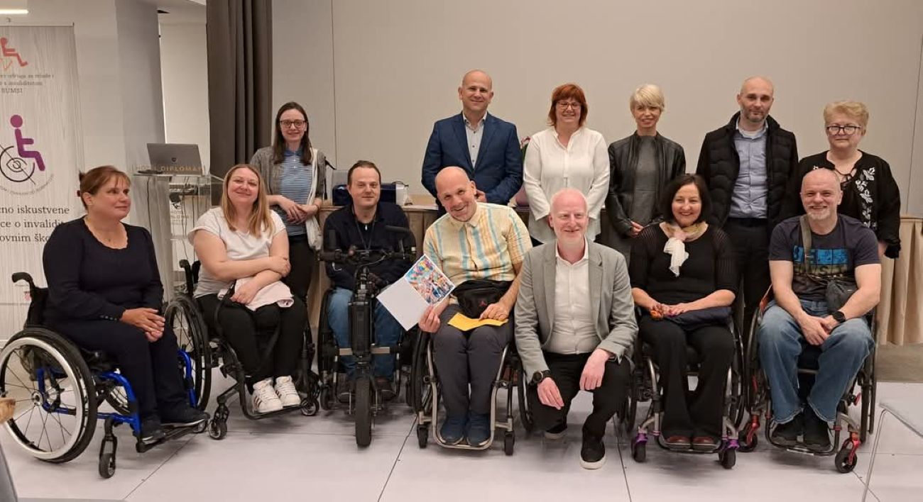 Na fotografiji je prikazana skupina ljudi okupljena u svečanom ozračju, vjerojatno povodom obilježavanja 16. rođendana Saveza SUMSI. Ukupno je četrnaest osoba, od kojih su neki u invalidskim kolicima, što odražava inkluzivnu prirodu događanja. Svi su nasmijani i opušteni, pozirajući za zajedničku fotografiju u dvorani, vjerojatno u hotelu Diplomat, gdje je i održana proslava. U pozadini se vidi plakat s logotipom i nazivom SUMSI-ja, dok je ispred njega postavljen govornik s natpisom hotela. Atmosfera je topla i prijateljska, a svi sudionici djeluju povezano i ponosno na zajednički rad i postignuća Saveza