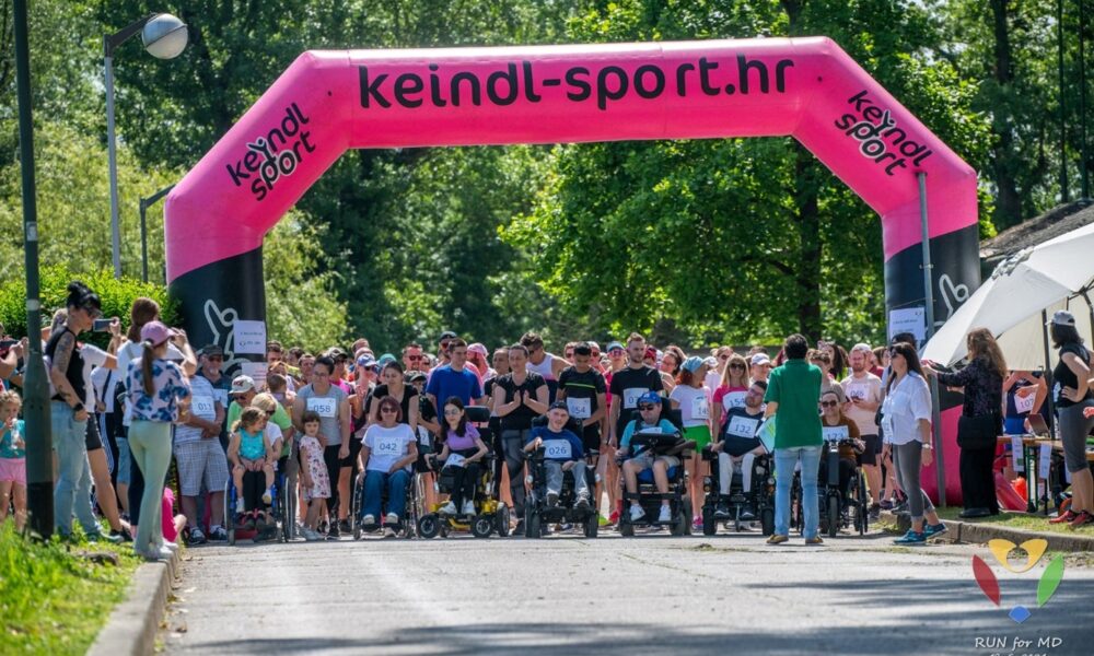 otografija prikazuje skupinu ljudi okupljenih ispod velikog ružičastog luka s natpisom „keindl-sport.hr“. Izgleda kao sportski događaj, jer mnogi sudionici nose brojeve na majicama. U prvom planu su osobe u invalidskim kolicima, što sugerira da je događaj inkluzivan i prilagođen osobama s invaliditetom. Pozadina je puna zelenila i drveća, što upućuje na to da se sve odvija na otvorenom, vjerojatno u parku ili nekom prirodnom okruženju. Na dnu desno nalazi se logo s natpisom „RUN for MD 19.5.2024.“, što sugerira da je događaj povezan s podizanjem svijesti ili prikupljanjem sredstava za mišićnu distrofiju. I