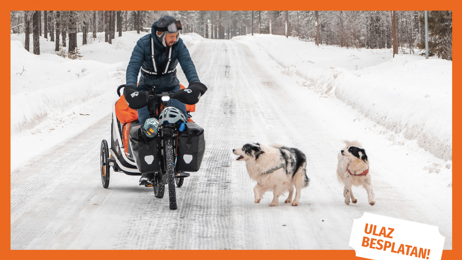 Muškarac odjeven u zimsku opremu vozi terenski bicikl s prikolicom po snježnoj cesti kroz šumu. Prate ga dva psa, jedan crno-bijeli i drugi bijeli s crnim mrljama. Snijeg prekriva cestu i okolicu, a prizor odaje dojam avanture i otpornosti. U donjem desnom kutu nalazi se narančasti natpis "ULAZ BESPLATAN!". Slika ima narančasti okvir.