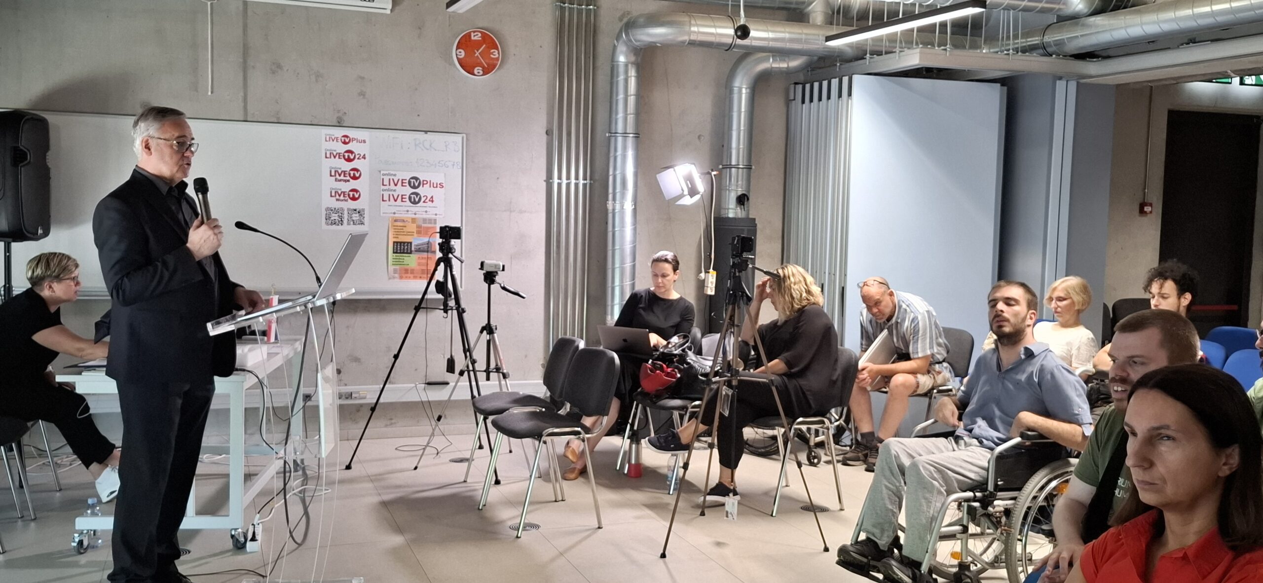 Fotografija prikazuje govornika u odjelu i publiku koja ga sluša.