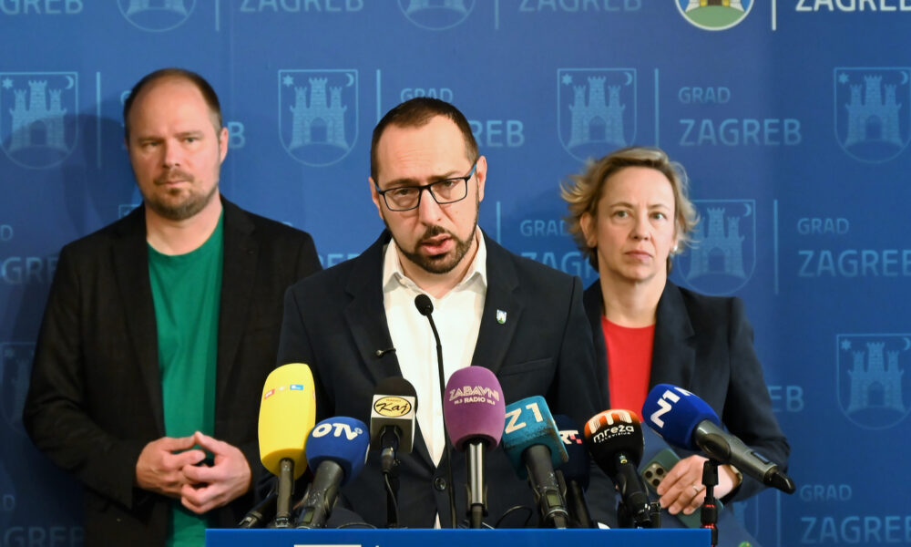 Gradonačelnik Zagreba stoji za govornicom i obraća se medijima, okružen brojnim mikrofonima različitih medijskih kuća. Iza njega stoje muškarac i žena ozbiljnog izraza lica. U pozadini je plavi zid s više puta ispisanim tekstom "GRAD ZAGREB" i stiliziranim grbom grada.