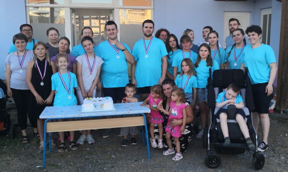 Na fotografiji je prikazana vesela skupina djece i odraslih okupljenih ispred zgrade, vjerojatno prostorija udruge Budi mi prijatelj iz Virovitice. Svi su okupljeni povodom proslave drugog rođendana udruge, što potvrđuje torta smještena na stolu ispred njih. Većina osoba nosi medalje oko vrata, što upućuje na sudjelovanje u sportskim ili rekreativnim aktivnostima koje su bile dio proslave. Grupa djeluje složno i radosno – djeca i odrasli su odjeveni u prepoznatljive plave majice, vjerojatno u znaku udruge, dok se nekoliko mlađe djece nalazi u prvim redovima, uz tortu. Desno se nalazi dječak u invalidskim kolicima, što dodatno potvrđuje inkluzivni karakter udruge. Ispred njega stoji žena koja drži malu djevojčicu u ružičastoj haljini. Atmosfera je prijateljska i svečana, a fotografija zrači zajedništvom, podrškom i pozitivnom energijom – u skladu s misijom udruge da djeci s teškoćama u razvoju i njihovim obiteljima pruži prostor za napredak, terapiju i međusobno povezivanje.