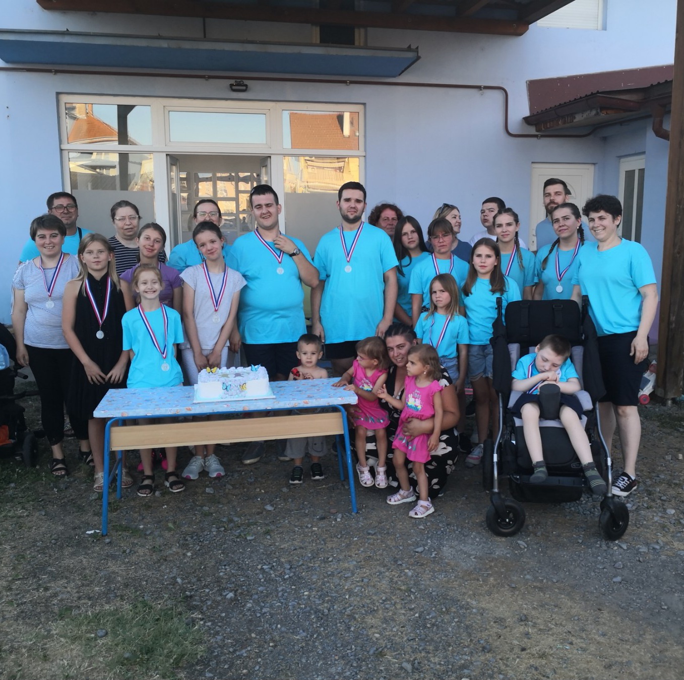 Na fotografiji je prikazana vesela skupina djece i odraslih okupljenih ispred zgrade, vjerojatno prostorija udruge Budi mi prijatelj iz Virovitice. Svi su okupljeni povodom proslave drugog rođendana udruge, što potvrđuje torta smještena na stolu ispred njih. Većina osoba nosi medalje oko vrata, što upućuje na sudjelovanje u sportskim ili rekreativnim aktivnostima koje su bile dio proslave. Grupa djeluje složno i radosno – djeca i odrasli su odjeveni u prepoznatljive plave majice, vjerojatno u znaku udruge, dok se nekoliko mlađe djece nalazi u prvim redovima, uz tortu. Desno se nalazi dječak u invalidskim kolicima, što dodatno potvrđuje inkluzivni karakter udruge. Ispred njega stoji žena koja drži malu djevojčicu u ružičastoj haljini. Atmosfera je prijateljska i svečana, a fotografija zrači zajedništvom, podrškom i pozitivnom energijom – u skladu s misijom udruge da djeci s teškoćama u razvoju i njihovim obiteljima pruži prostor za napredak, terapiju i međusobno povezivanje.