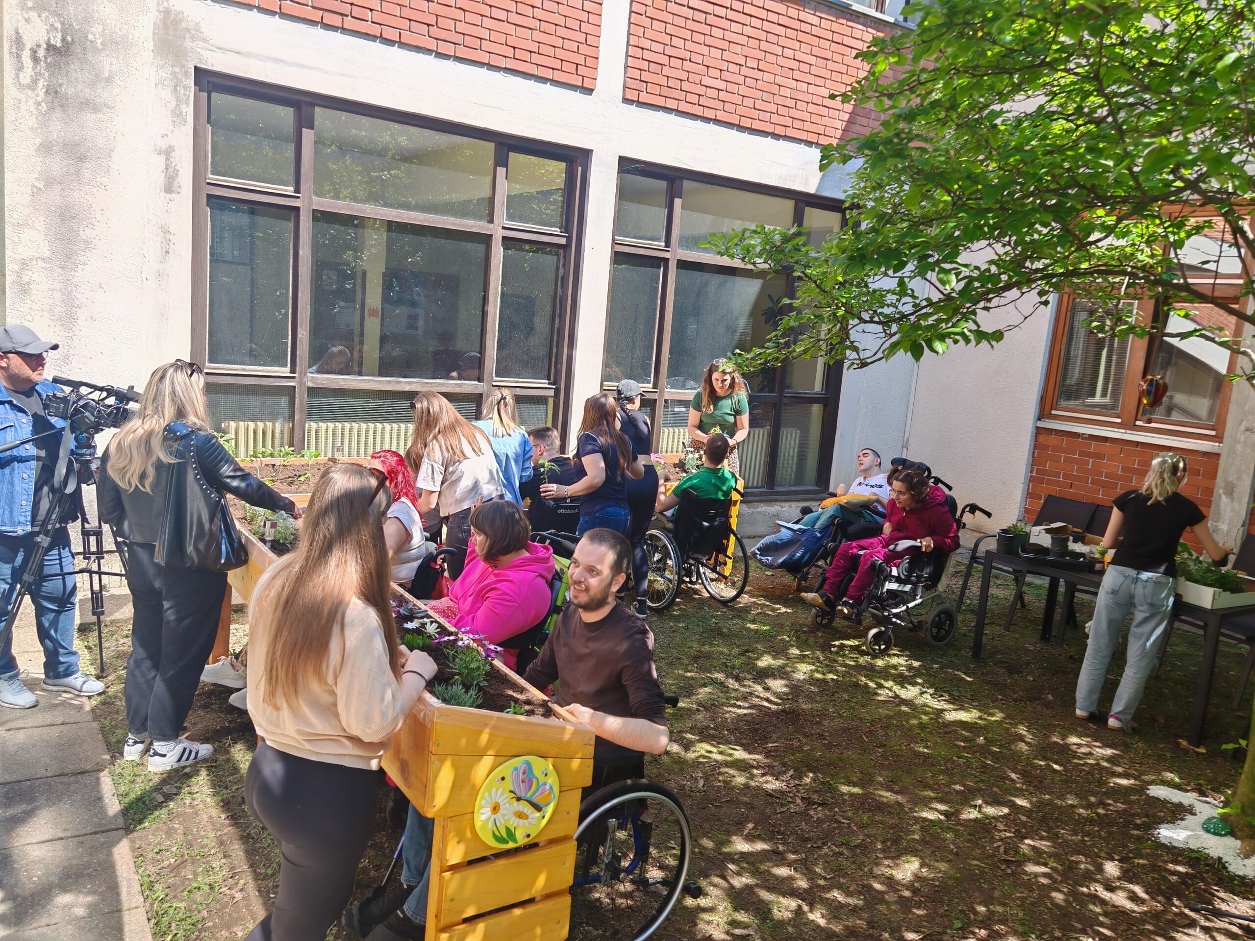 Na fotografiji je prikazana živa i vedra scena u dvorištu Centra za pružanje usluga u zajednici Varaždin. Grupa korisnika i djelatnika Centra sudjeluje u vrtnoj radionici na otvorenom. U prvom planu vidimo podignutu vrtnu gredicu prilagođenu osobama u invalidskim kolicima, oko koje su okupljeni sudionici – neki sjede u kolicima, dok drugi stoje i pomažu pri sadnji biljaka. Atmosfera je topla i inkluzivna, a svi prisutni djeluju angažirano i nasmijano. U pozadini se nalaze velika staklena okna zgrade Centra, a oko sudionika se širi zelenilo – drveće i trava stvaraju ugodno, prirodno okruženje. S lijeve strane u kadar ulazi snimateljska ekipa s kamerama, što sugerira da se događaj medijski prati. Na desnoj strani kadra još nekoliko sudionika uređuje biljke na stolu, što dodatno naglašava radnu i zajedničku atmosferu. Ova fotografija prikazuje primjer dobre prakse socijalne inkluzije, zajedništva i terapijskog rada kroz aktivnosti u prirodi, koje doprinose dobrobiti i kvaliteti života korisnika Centra.