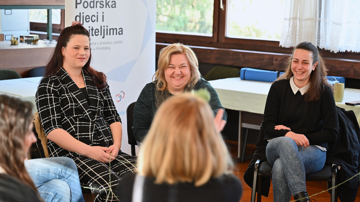 Tri žene sjede u krugu i razgovaraju, nasmijane i opuštene, u prostoriji za edukacije. U pozadini je roll-up plakat s natpisom "Podrška djeci i roditeljima", koji sugerira da je riječ o radionici ili edukaciji vezanoj uz psihosocijalnu podršku. Oko njih se nalaze drugi sudionici, djelomično vidljivi, a prostor je svijetao i ugodan, s drvenim okvirima prozora i stolovima u pozadini. Atmosfera je topla i podržavajuća.