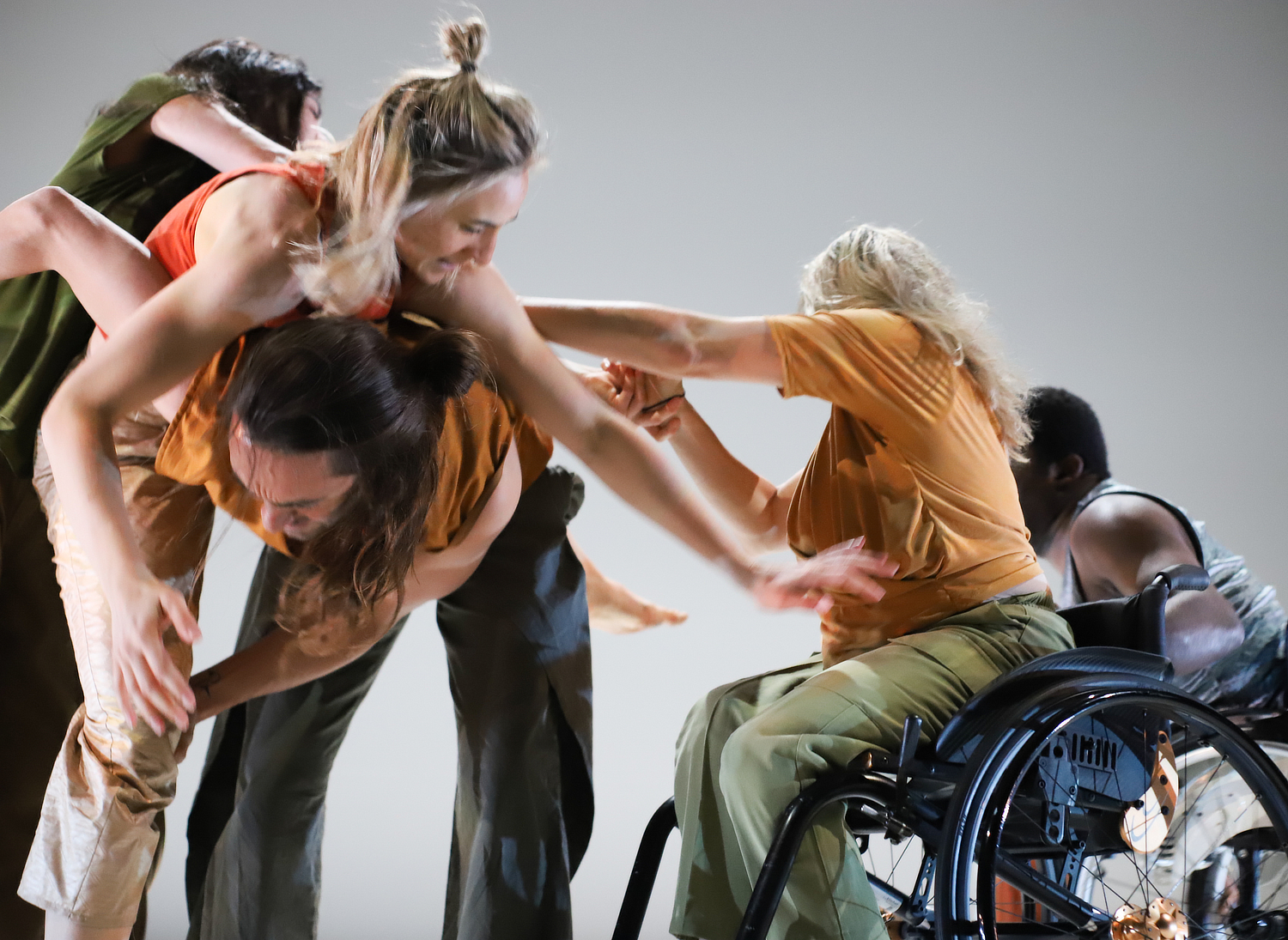Grupa plesača u intenzivnom pokretu izvodi suvremeni ples. Među njima je žena u invalidskim kolicima koja aktivno sudjeluje, pružajući ruku prema ostalim izvođačima. Atmosfera je energična i puna pokreta, s izraženom tjelesnom interakcijom i izražajnim pokretima tijela.