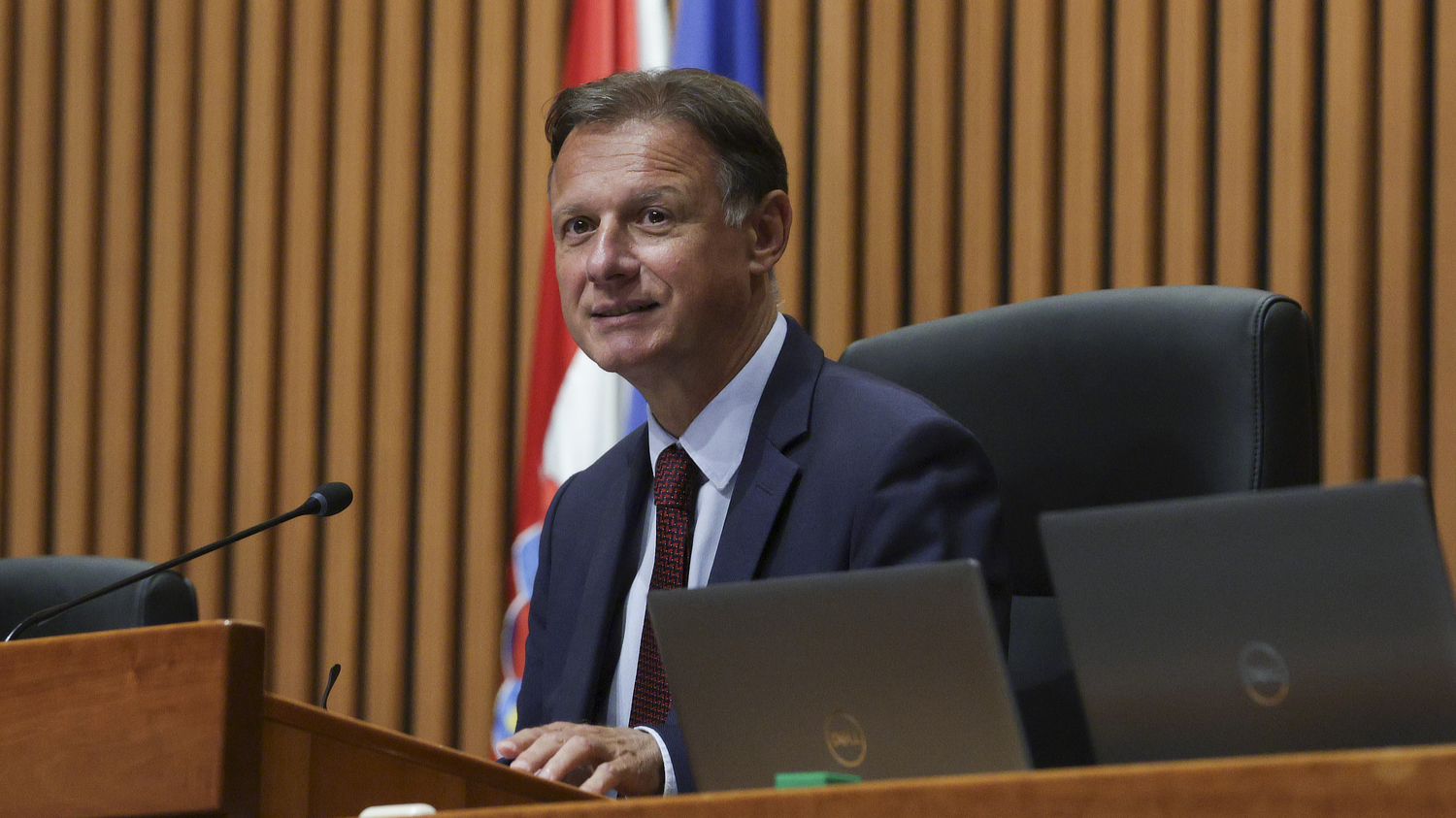 Muškarac u odijelu i kravati sjedi za govornicom u velikoj dvorani, s hrvatskom i europskom zastavom u pozadini. Ispred njega su dva prijenosna računala, a izražava lagani osmijeh.