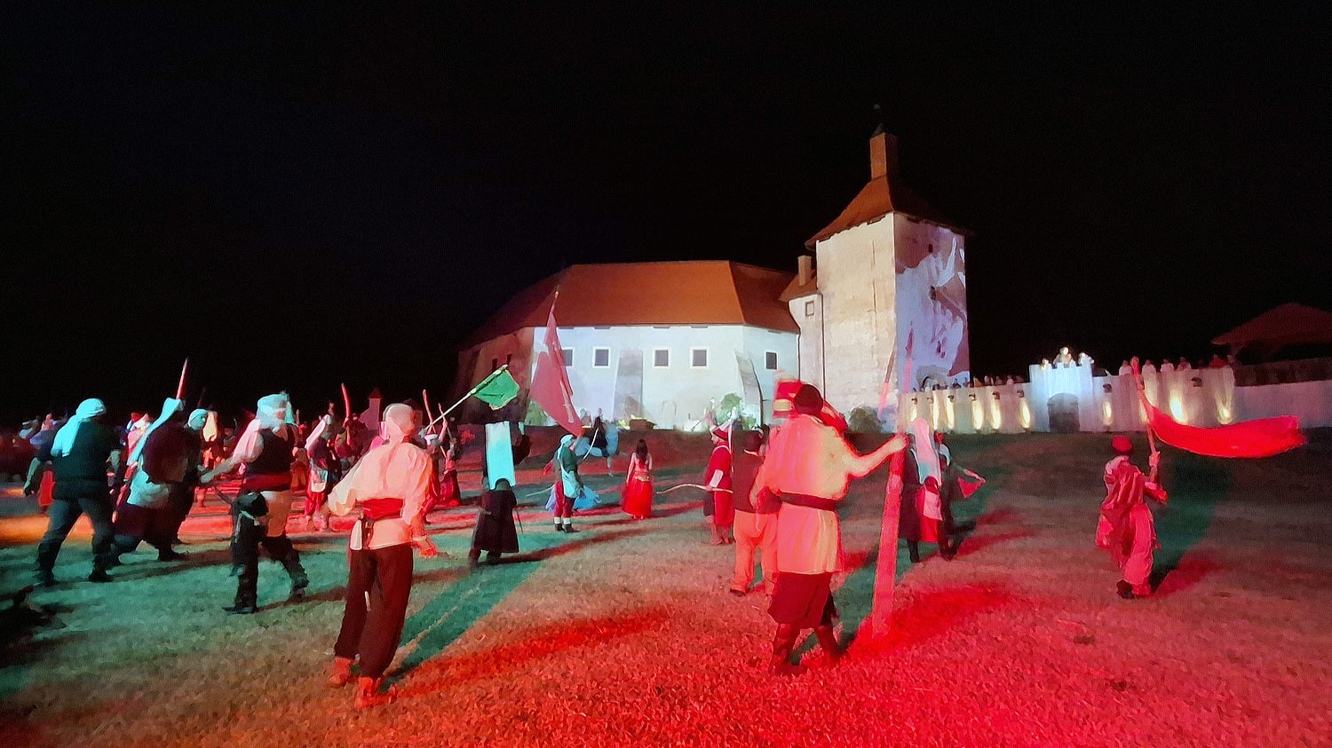 Noćna scena povijesne rekonstrukcije ispred utvrde. Brojni sudionici u srednjovjekovnim kostimima, s mačevima i zastavama, izvode koreografiju pod obojenim svjetlima. U pozadini je osvijetljena kamena kula i dio publike.