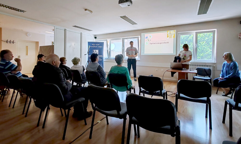 Manja dvorana s nekoliko redova stolica na kojima sjede starije osobe, usmjerene prema projekcijskom platnu. Na platnu piše „Optimizam prošlosti“, uz ilustraciju osobe i dekorativne cvjetove. Dvoje predavača stoji ispred platna, a treća osoba sjedi sa strane. U prostoru vlada tiha, edukativna atmosfera. Na zidu se vidi plakat s porukom: „Nije svaki invaliditet vidljiv“.