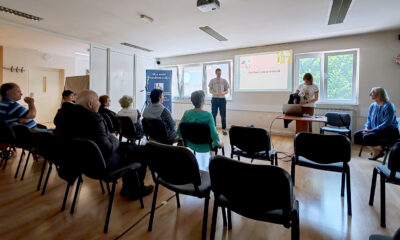 Manja dvorana s nekoliko redova stolica na kojima sjede starije osobe, usmjerene prema projekcijskom platnu. Na platnu piše „Optimizam prošlosti“, uz ilustraciju osobe i dekorativne cvjetove. Dvoje predavača stoji ispred platna, a treća osoba sjedi sa strane. U prostoru vlada tiha, edukativna atmosfera. Na zidu se vidi plakat s porukom: „Nije svaki invaliditet vidljiv“.