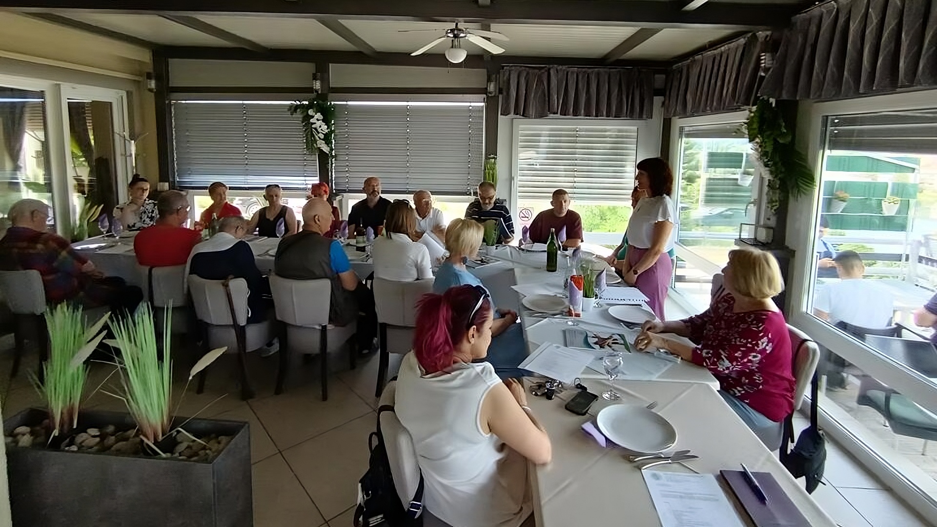 Skupina odraslih osoba sjedi oko dugog stola u zatvorenom dijelu restorana. Jedna žena stoji i obraća se grupi, dok ostali slušaju. Na stolu su postavljeni tanjuri, čaše i bilješke. Atmosfera je radna, ali neformalna, a prostor je prozračan s mnogo prirodnog svjetla i ukrasnim biljkama.