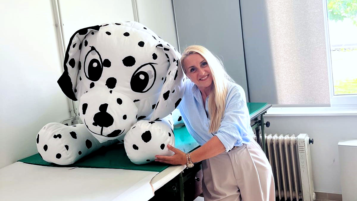 Žena svijetle kose, odjevena u svijetloplavu košulju i svijetle hlače, nasmiješeno pozira pored velikog plišanog dalmatinera s crnim točkicama. Nalaze se u svijetloj prostoriji uz prozor i radijator.