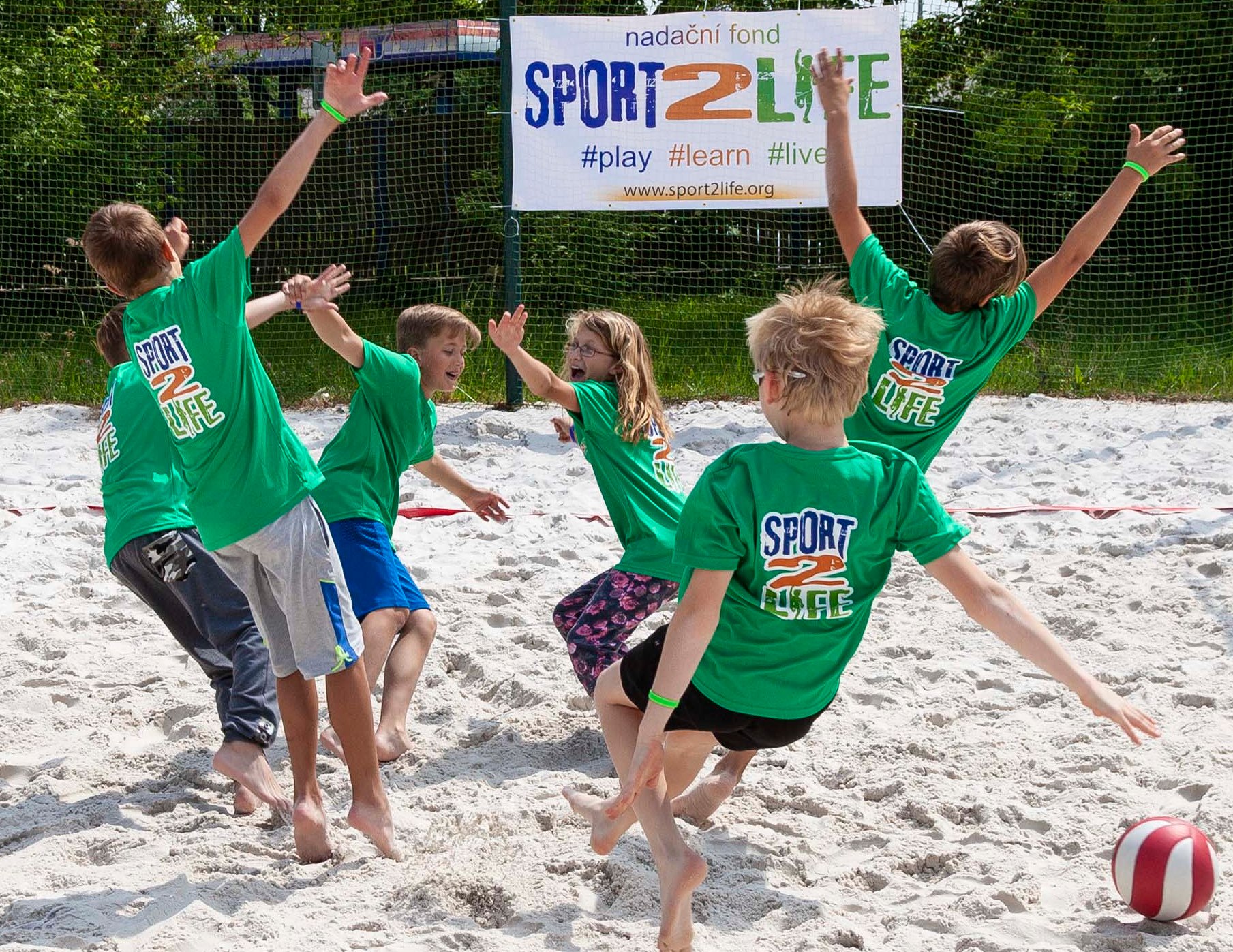 Fotografija prikazuje djecu u zelenim dresovima kako padaju po pijsku, dok na bijelom plakatu piše ime sportskog događaja 'SPORT2LIFE'.