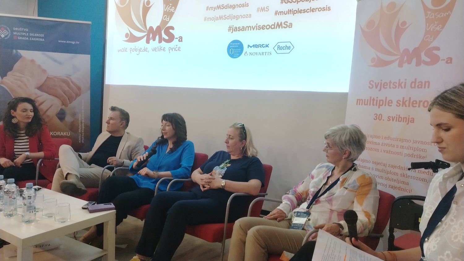 Panel rasprava povodom Svjetskog dana multiple skleroze. Šest sudionika, uključujući muškarce i žene, sjedi u nizu na pozornici ispred projekcije s logom kampanje "Ja sam više od MS-a". U pozadini su promotivni plakati s natpisima poput „male pobjede, velike priče“, „Svjetski dan multiple skleroze – 30. svibnja“, te hashtagovi povezani s dijagnozom MS-a. Atmosfera je ozbiljna i informativna.