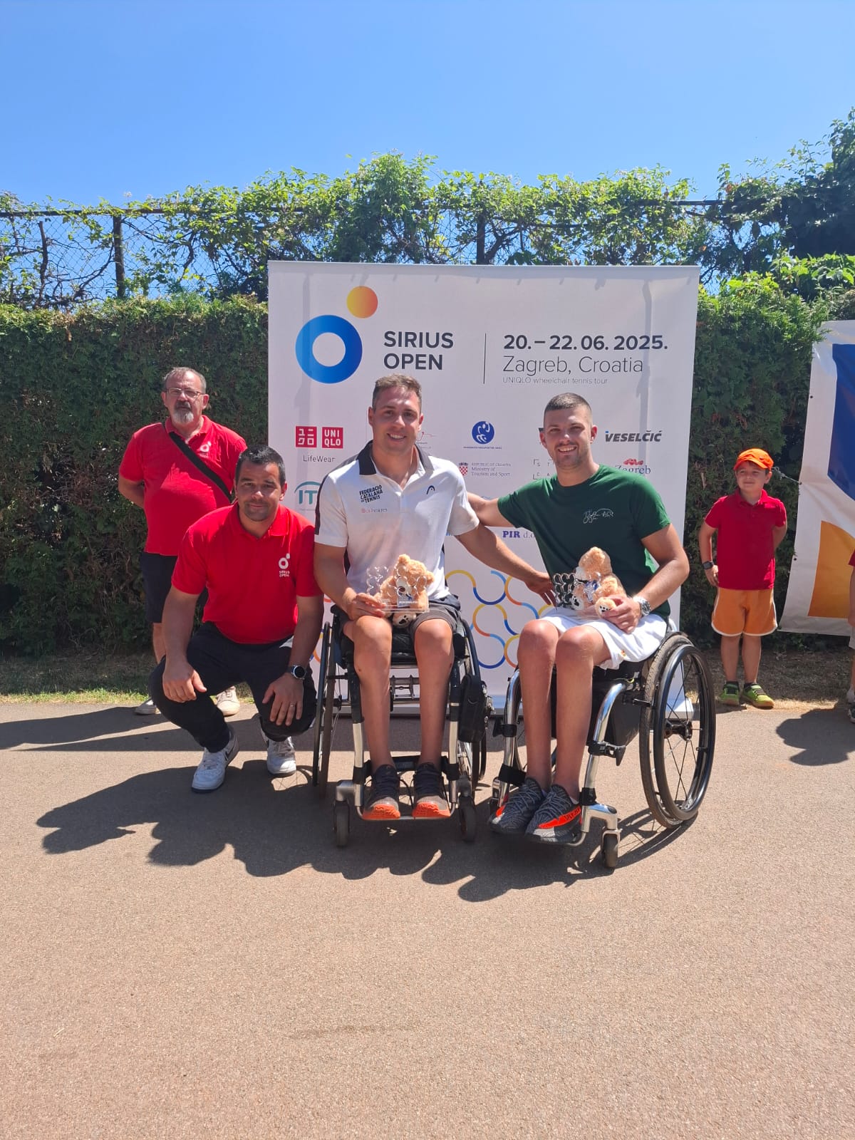 Fotografija prikazuje tri osobe, dvoje je u kolicima i drži magrade te plišane medvjediće, dok treća osoba čuči pokraj njih.