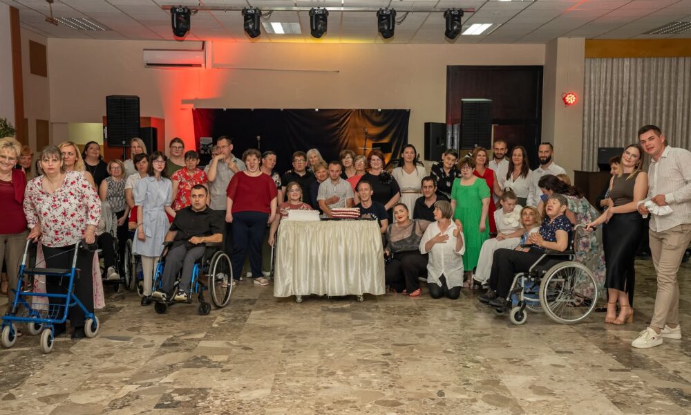 Na fotografiji je prikazana grupa ljudi okupljena u zatvorenom prostoru. Neki od njih stoje, dok drugi sjede u invalidskim kolicima ili koriste hodalice. U središtu slike nalazi se stol prekriven bijelim stolnjakom, na kojem su postavljene dvije torte. Iza grupe ljudi vidi se pozornica s crnim zastorom i nekoliko zvučnika. Prostor je osvijetljen reflektorima, a na stropu su dodatna rasvjetna tijela. Pod je prekriven pločicama u nijansama smeđe boje. Fotografija odaje dojam proslave ili okupljanja, pri čemu se ističe raznolikost ljudi u pogledu dobi i fizičkih sposobnosti.