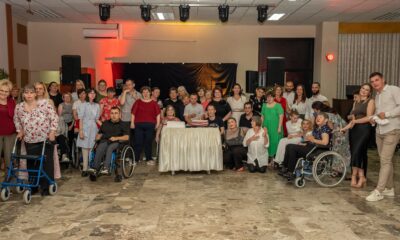 Na fotografiji je prikazana grupa ljudi okupljena u zatvorenom prostoru. Neki od njih stoje, dok drugi sjede u invalidskim kolicima ili koriste hodalice. U središtu slike nalazi se stol prekriven bijelim stolnjakom, na kojem su postavljene dvije torte. Iza grupe ljudi vidi se pozornica s crnim zastorom i nekoliko zvučnika. Prostor je osvijetljen reflektorima, a na stropu su dodatna rasvjetna tijela. Pod je prekriven pločicama u nijansama smeđe boje. Fotografija odaje dojam proslave ili okupljanja, pri čemu se ističe raznolikost ljudi u pogledu dobi i fizičkih sposobnosti.