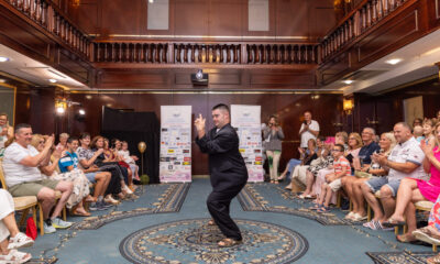 Mlađi muškarac u odijelu s Down sindromom samouvjereno pozira i pleše na pisti modne revije. Oko njega sjedi publika koja plješće i veselo gleda izvedbu. Prostor je elegantan, s tepisima i drvenim zidovima, a u pozadini se vide sponzorski plakati.