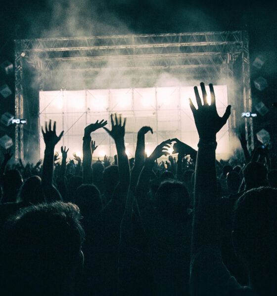 Noćna scena velikog koncerta na otvorenom. Mnoštvo ljudi u publici drži ruke u zraku, dok je pozornica u pozadini obasjana snažnim svjetlima i okružena dimnim efektima. Na obje strane pozornice nalaze se veliki ekrani s projekcijama. Atmosfera je energična, dinamična i ispunjena uzbuđenjem.