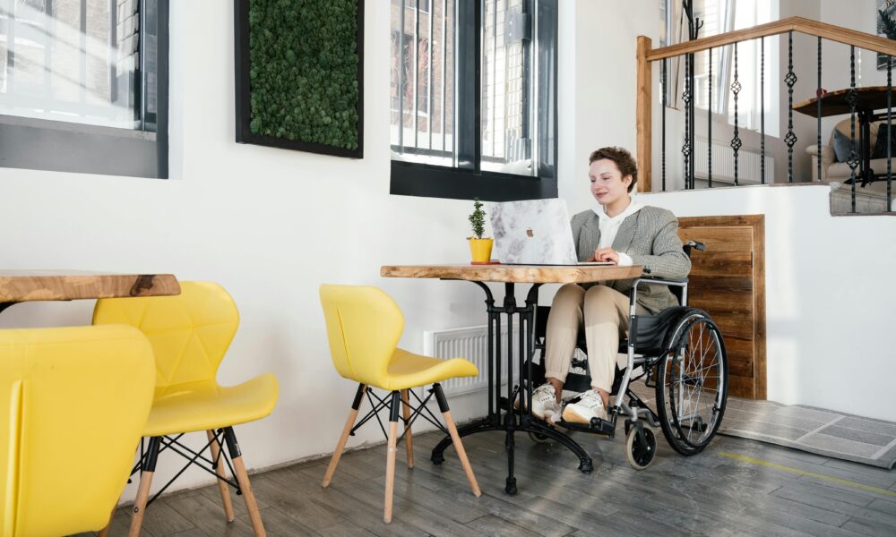 Na slici je osoba koja sjedi ui invalidskim kolicima i koristi laptop