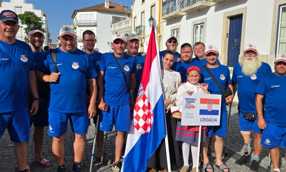 Slika prikazuje više ljudi u plavim majicama. Ispred njih su dvije osoba u bijelim majicama, jedna drži natpis na kojem piše 'Croacia' i nacrtana je hrvatska zastava, dok druga drži hrvatsku zastavu. Neki ljudi na slici umjesto noge imaju protezu.