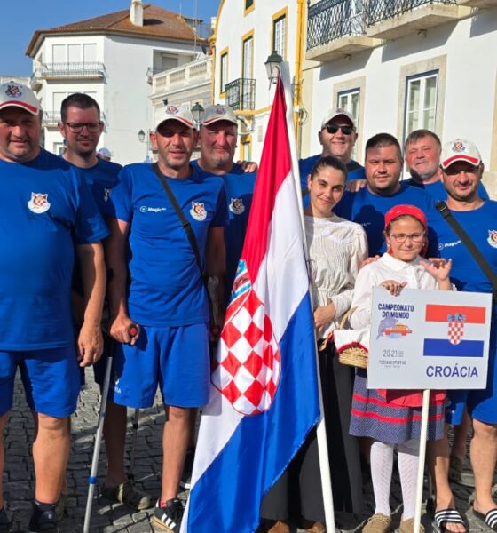 Slika prikazuje više ljudi u plavim majicama. Ispred njih su dvije osoba u bijelim majicama, jedna drži natpis na kojem piše 'Croacia' i nacrtana je hrvatska zastava, dok druga drži hrvatsku zastavu. Neki ljudi na slici umjesto noge imaju protezu.