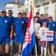 Slika prikazuje više ljudi u plavim majicama. Ispred njih su dvije osoba u bijelim majicama, jedna drži natpis na kojem piše 'Croacia' i nacrtana je hrvatska zastava, dok druga drži hrvatsku zastavu. Neki ljudi na slici umjesto noge imaju protezu.